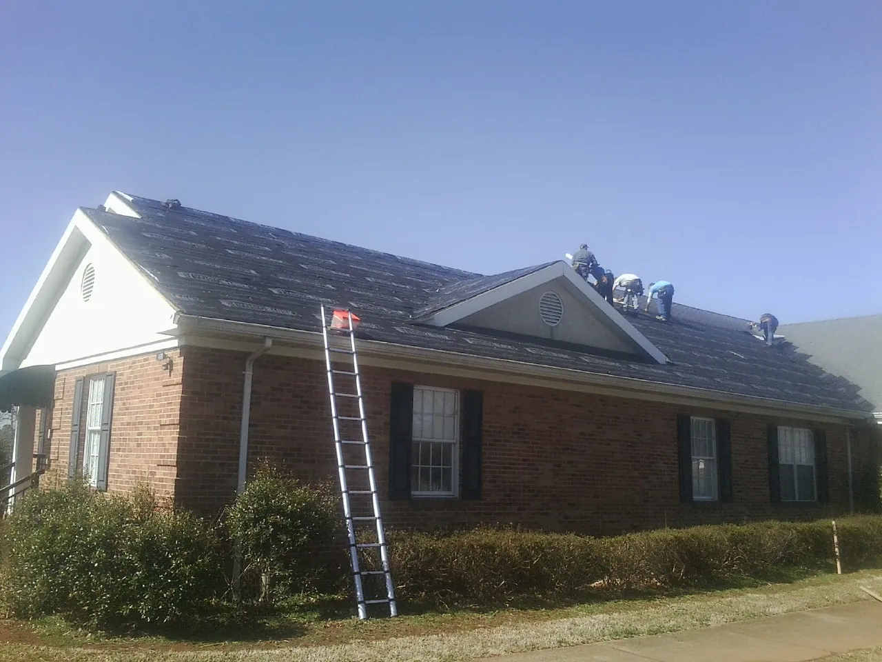 Crew tearing shingles off a brick ranch roof with a ladder set — Metal Roof Repair work by our crew in Clio