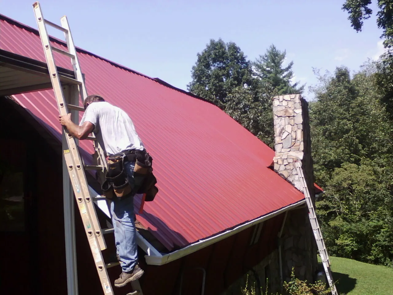 Roofer on a ladder beside a red metal roof and stone chimney — Soffit & Fascia Repair by Roofing Brothers in Clio